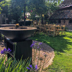 Bol en métal noir avec un rebord en bois et des bûches empilées à l’intérieur, reposant sur une base en piédestal noir assortie.