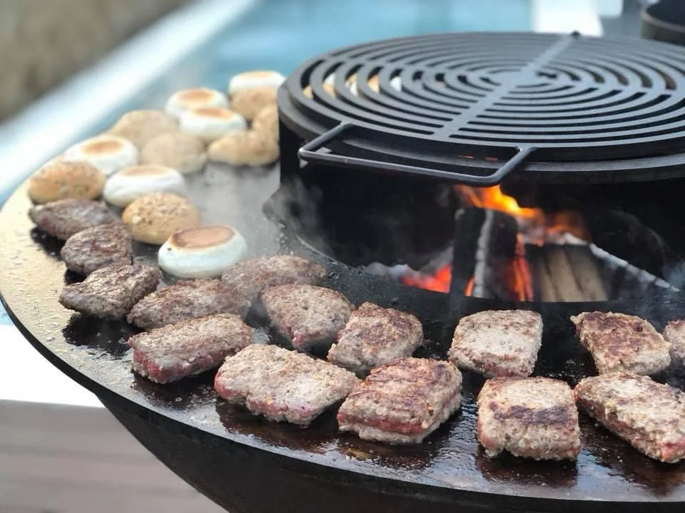 Gril en fonte noire avec une grille circulaire et des braises incandescentes en dessous, cuisant des galettes de viande assaisonnées à côté de guimauves grillées.