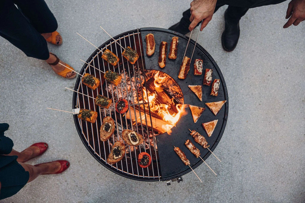 Foyer de feu rond pour grill avec grille métallique et bûches de bois en train de brûler à l’intérieur, présentant des brochettes de viande et de légumes.