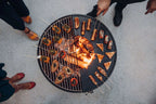 Foyer de feu rond pour grill avec grille métallique et bûches de bois en train de brûler à l’intérieur, présentant des brochettes de viande et de légumes.