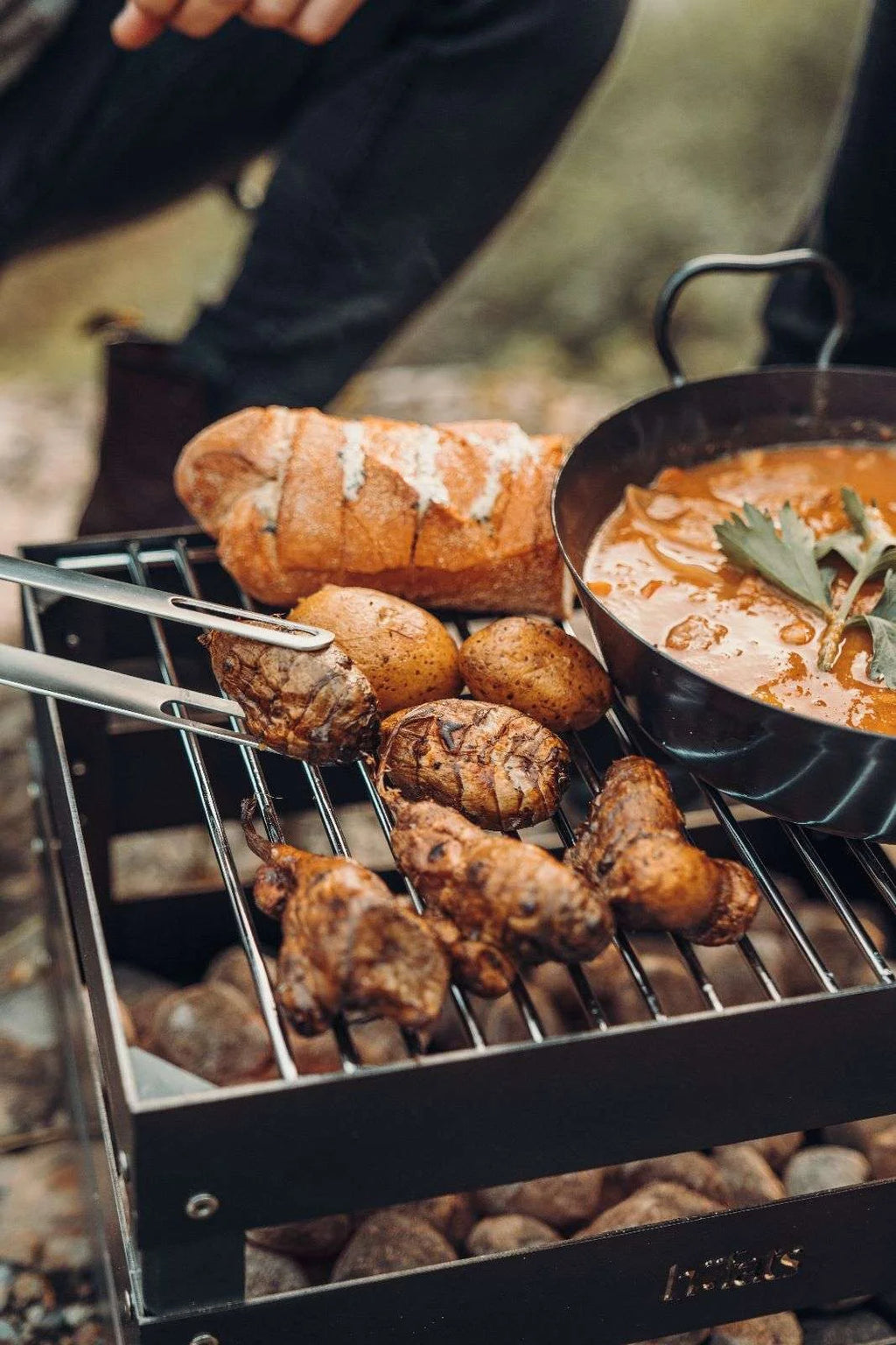 Barbecue au charbon avec des ailes de poulet et des pommes de terre sur la grille, accompagnée d’une casserole de soupe.
