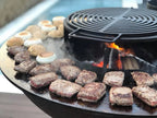 Gril en fonte noire avec une grille circulaire et des braises incandescentes en dessous, cuisant des galettes de viande assaisonnées aux côtés de guimauves grillées.