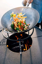 Wok noir avec des légumes colorés et une poignée en bois pour remuer, posé sur un support de feu de charbon.