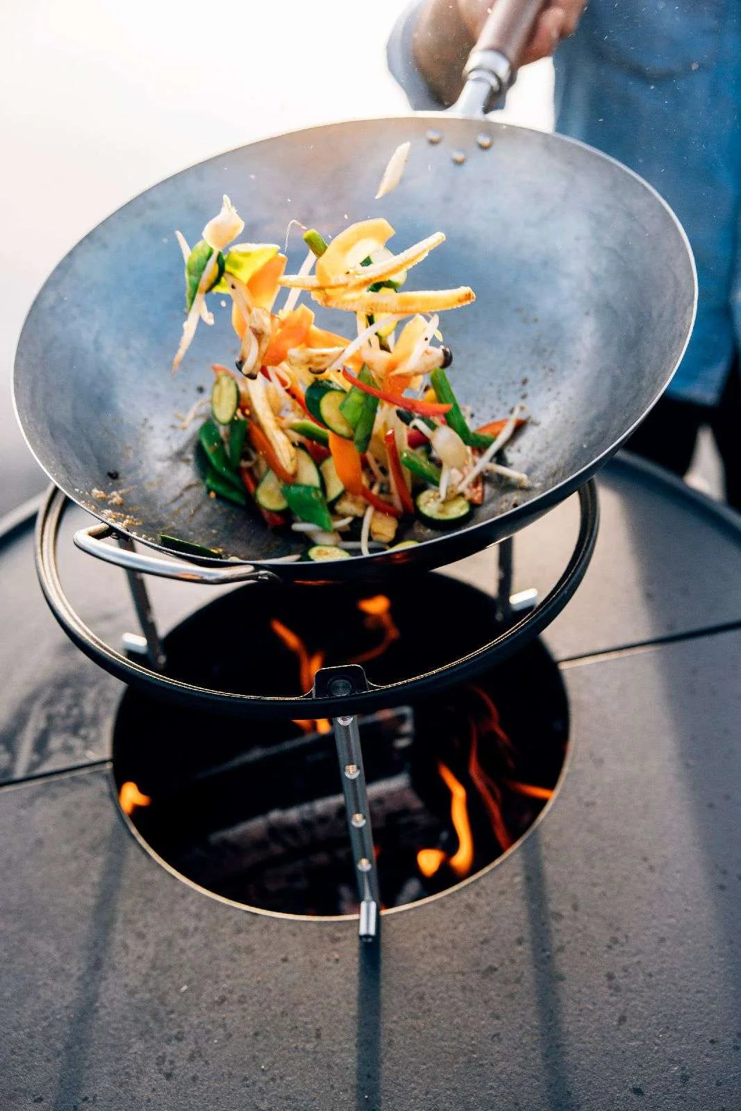 Wok noir avec des légumes colorés et une poignée en bois pour remuer, posé sur un support de feu de charbon.
