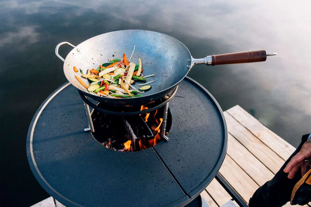 Wok noir avec manche en bois et rebord en métal, cuisant sur feu ouvert sur le quai.
