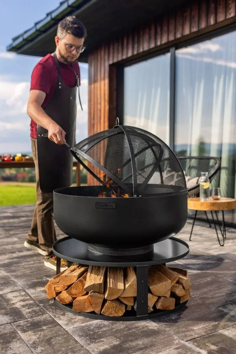 Foyer de feu noir avec un écran en maille et un support à bois, conçu pour un usage extérieur.