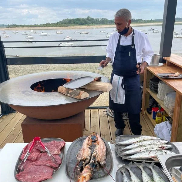 Un chef portant une chemise blanche et un tablier sombre se tient à côté d’un grand foyer métallique rond avec des flammes à l’intérieur.