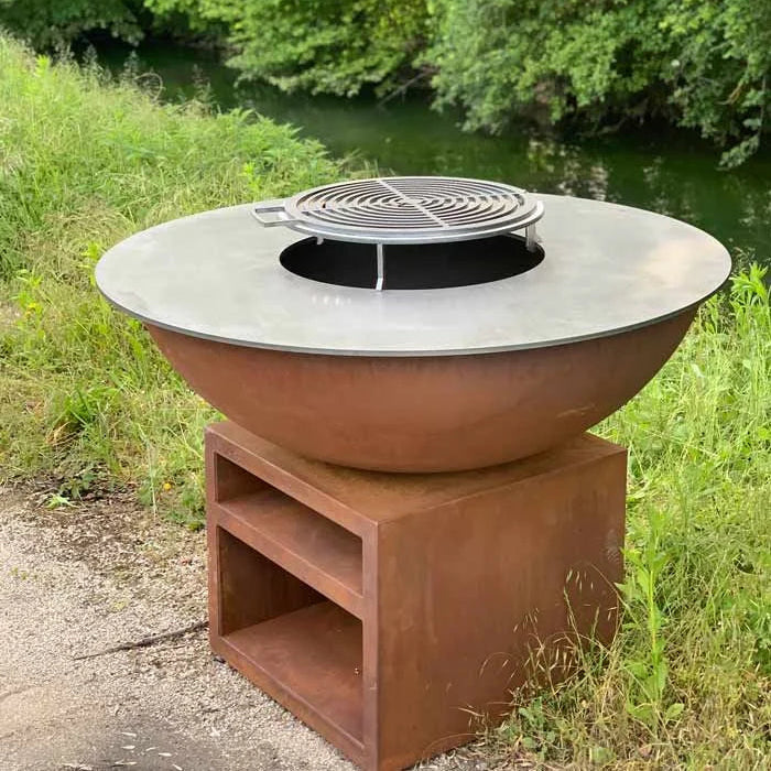 Foyer de feu rond rustique avec un dessus métallique plat et une grille de gril intégrée, reposant sur une base en acier rouillé assortie en forme de piédestal.