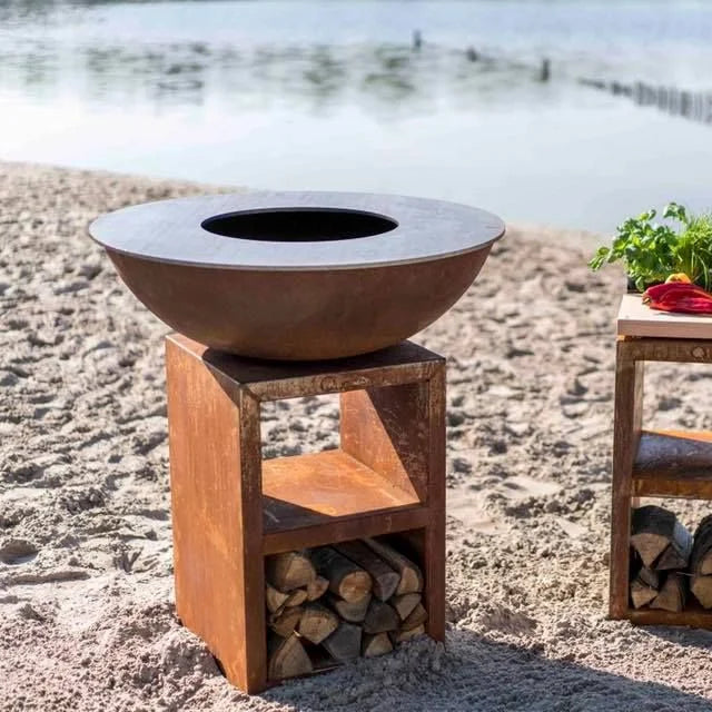 Bol en acier de couleur cuivre rouillé avec une ouverture circulaire, reposant sur un support carré assorti qui retient des bûches empilées en dessous.