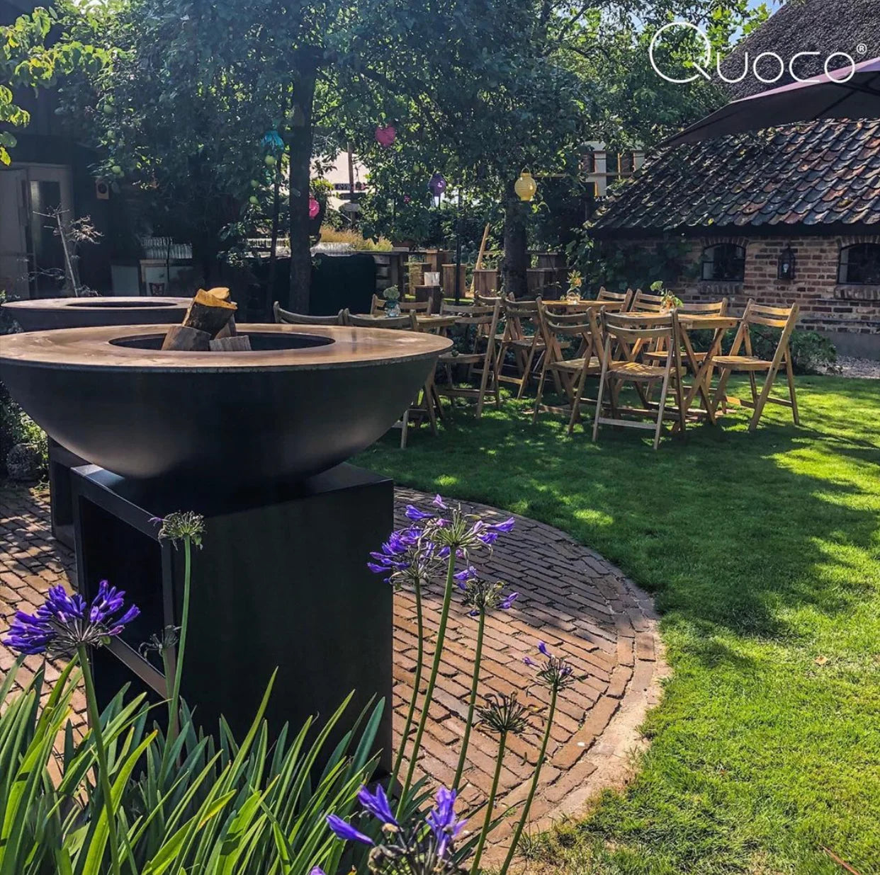 Bol en métal noir avec un rebord en bois et des bûches empilées à l’intérieur, reposant sur une base en piédestal noir assortie.