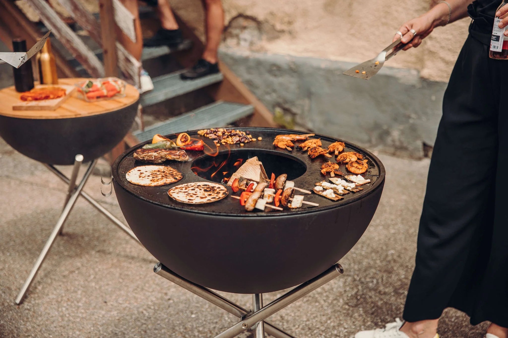 Grill barbecue rond noir avec pieds et poignée en acier inoxydable, présentant une variété d’aliments sur la surface de cuisson.