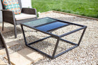 Table basse d’extérieur à cadre en métal noir avec un plateau en verre comportant deux sections distinctes : une claire et une foncée, installée sur une terrasse en gravier.