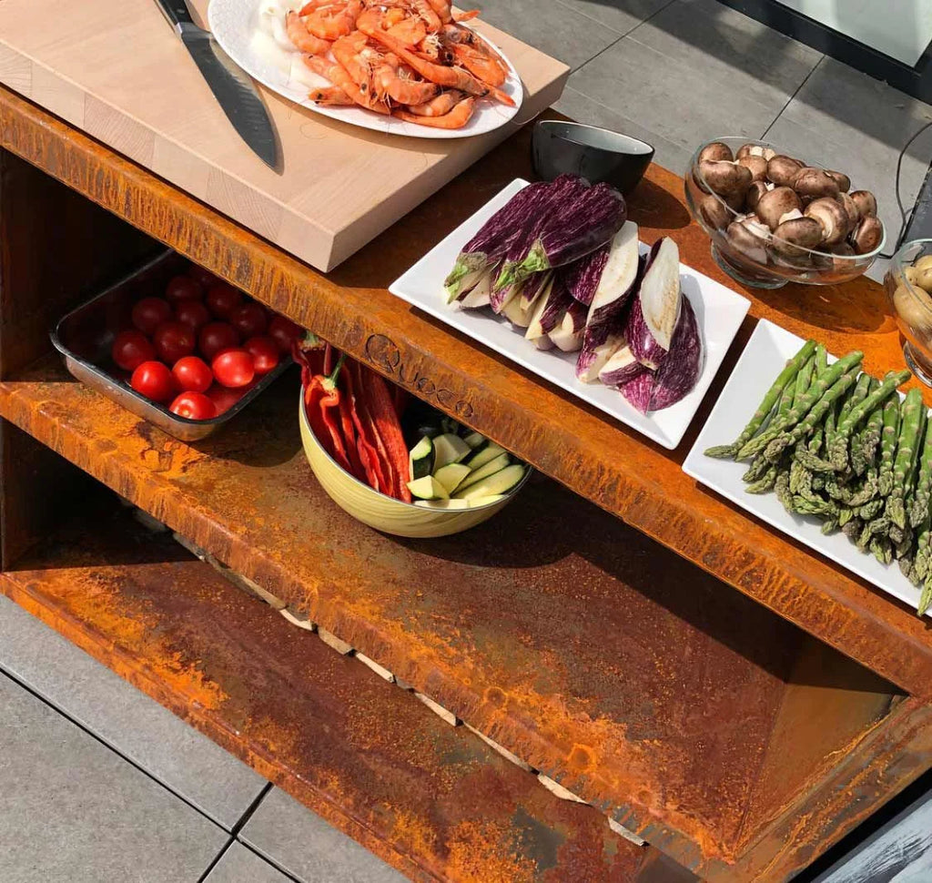 Îlot de cuisine en métal rouillé orange avec étagères ouvertes contenant des produits frais et une assiette de crevettes cuites