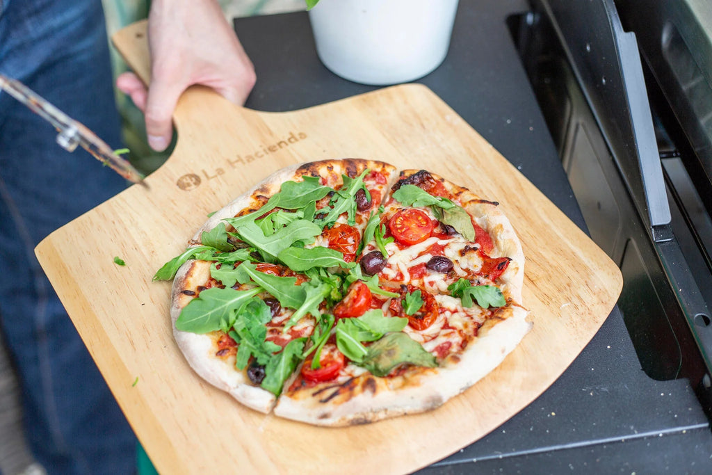 Une pizza fraîchement cuite, garnie de roquette, de tomates cerises et d’olives, repose sur une planche de service en bois clair.
