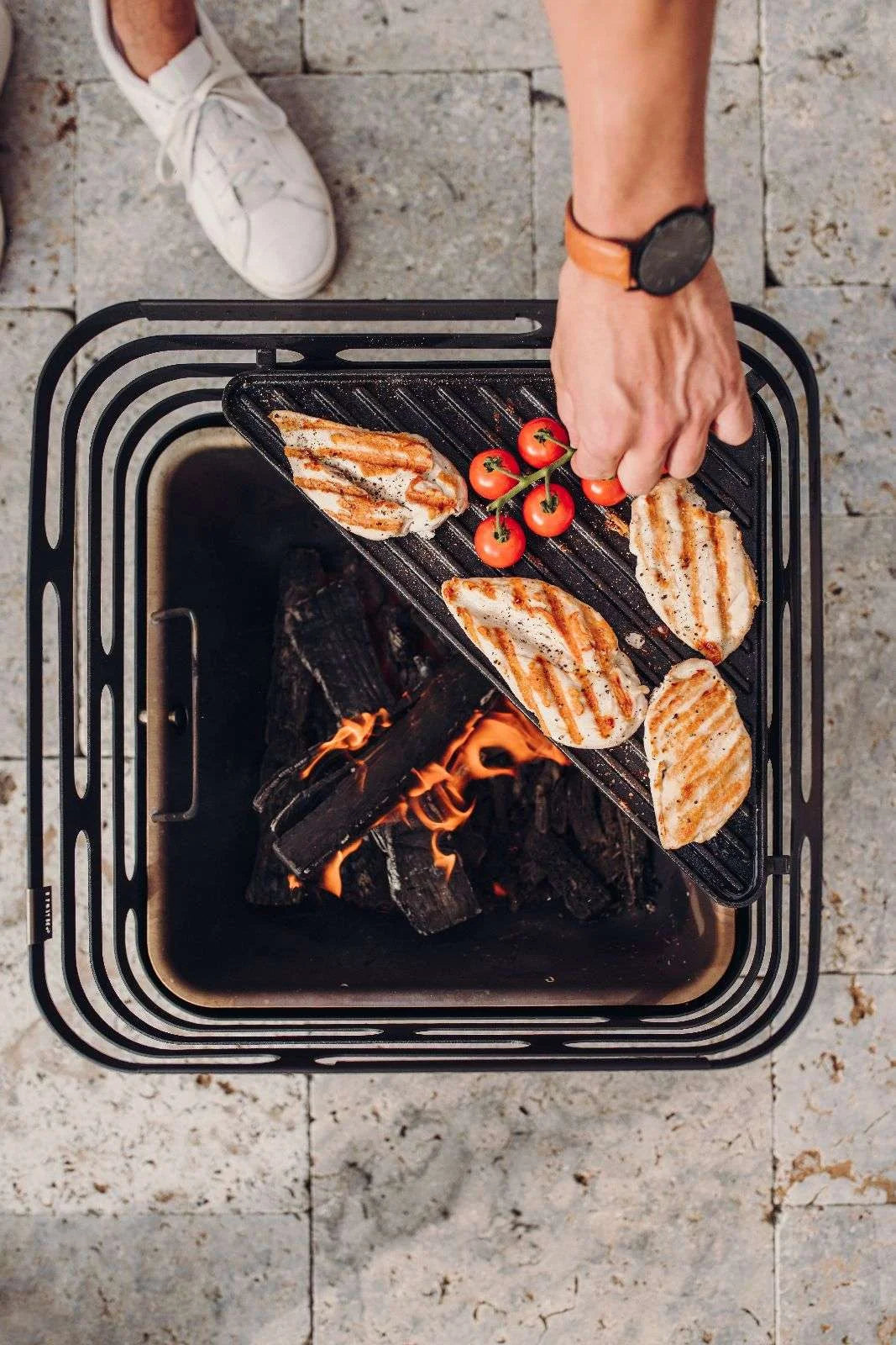 Barbecue au charbon avec grille noire et charbons rouges, baskets blanches visibles en arrière-plan.