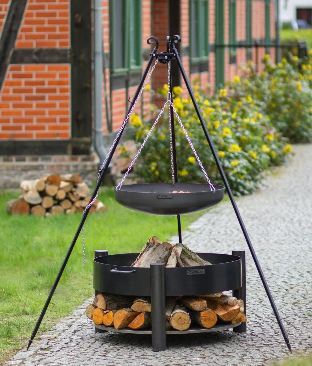 Foyer de feu en métal noir avec gril suspendu et bûches pour cuisiner en extérieur.