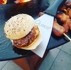 Un burger de bœuf juteux avec un pain grillé parsemé de graines repose sur un plateau en papier au-dessus d’une spatule marquée ’quoco’.