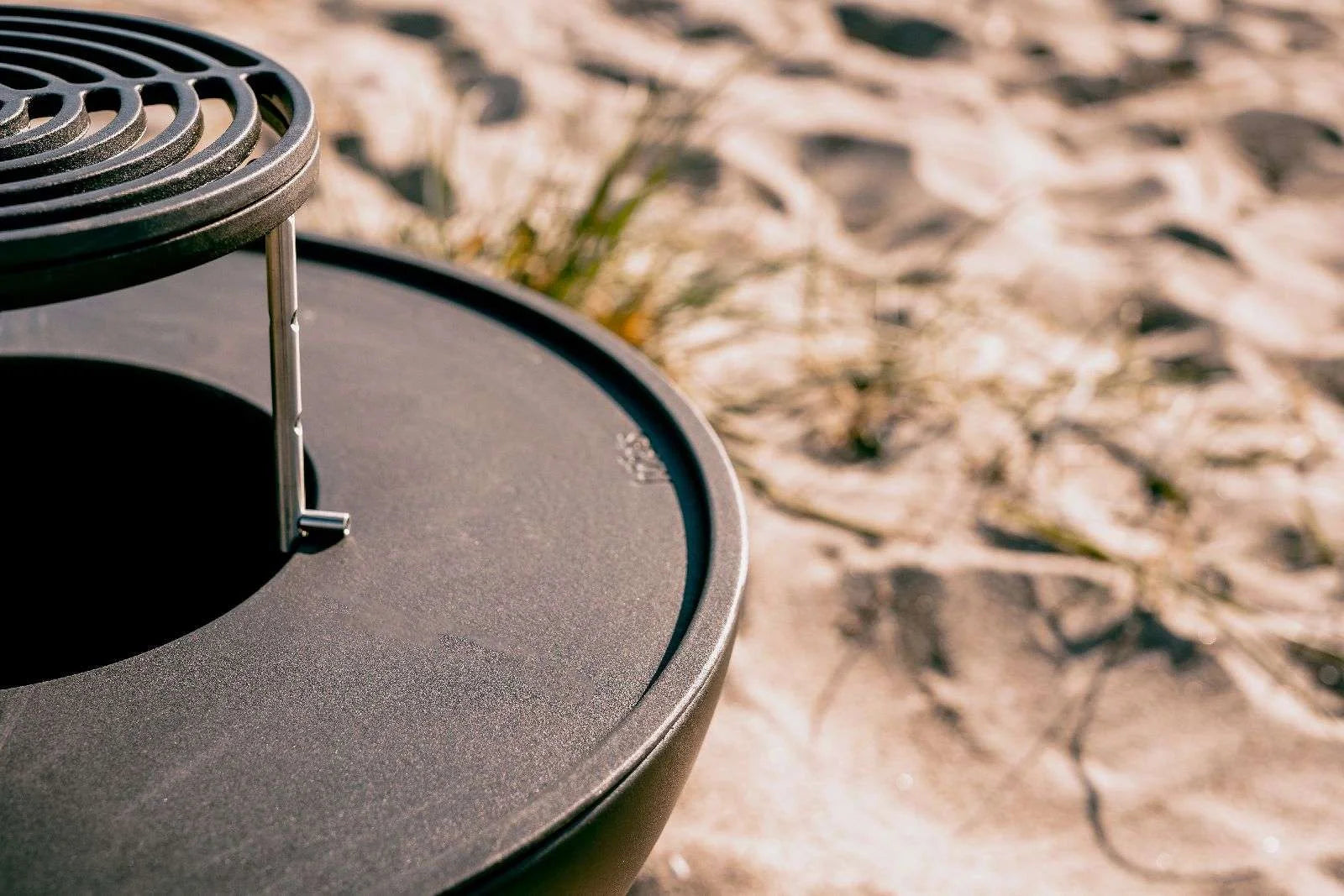 Gril rond noir avec grille et poignée sur fond de sable de plage.