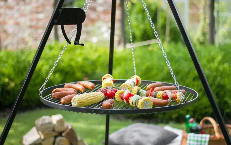 Gril suspendu avec cadre en métal noir et chaînes, contenant divers aliments comme des saucisses, du maïs en épi, des brochettes.
